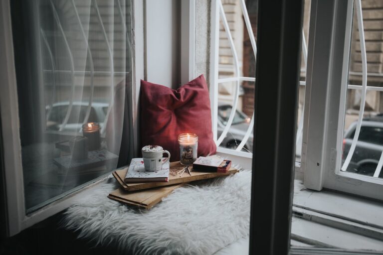A hygge setup of a red pillow, a lit candle, a mug of hot cocoa and a fuzzy blanket nestled in a window seat