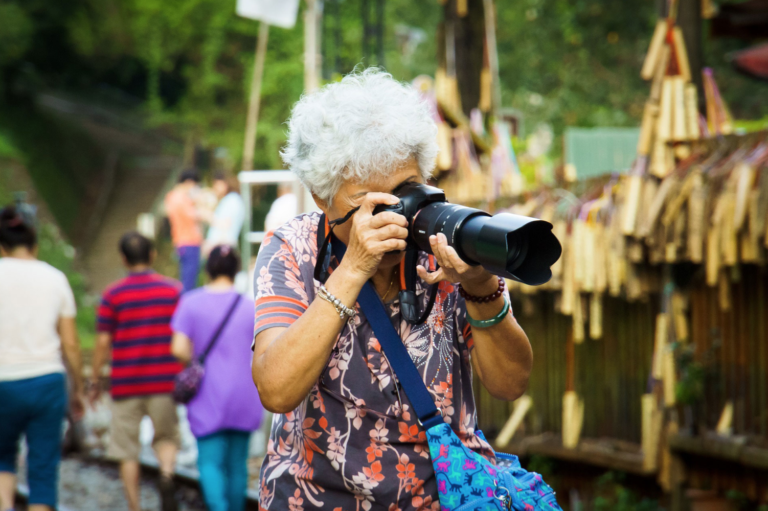 An older adult woman holds a DSLR camera with a hooded zoom lens up to her face, in the act of taking a photo